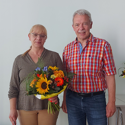Middle-aged woman holding a colorful bouquet of flowers standing next to a smiling middle-aged man in a red plaid shirt against a plain background.