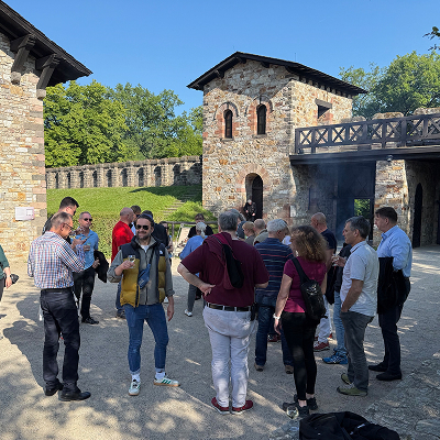 Group of people gathered outdoors near a stone building and bridge on a sunny day.