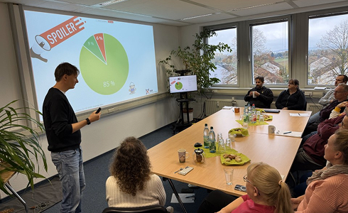 A young man presenting a pie chart labeled 'Spoiler' to a group seated around a conference table with drinks and snacks in a well-lit office room.