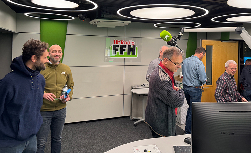 Five men standing and conversing inside a radio studio with a Hit Radio FFH logo on the wall and microphones overhead.